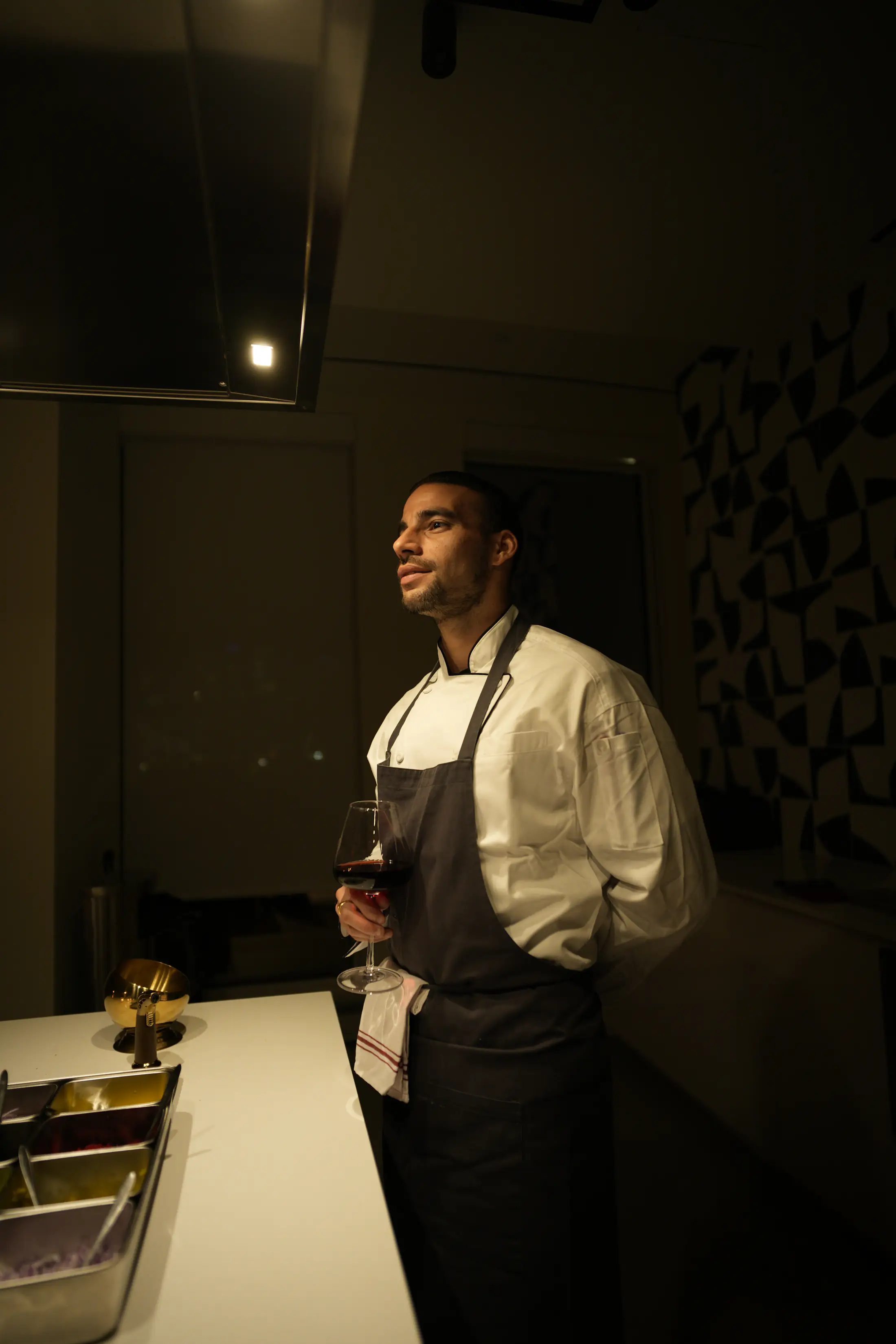 A chef in whites and a dark apron holding a glass of red wine in the kitchen