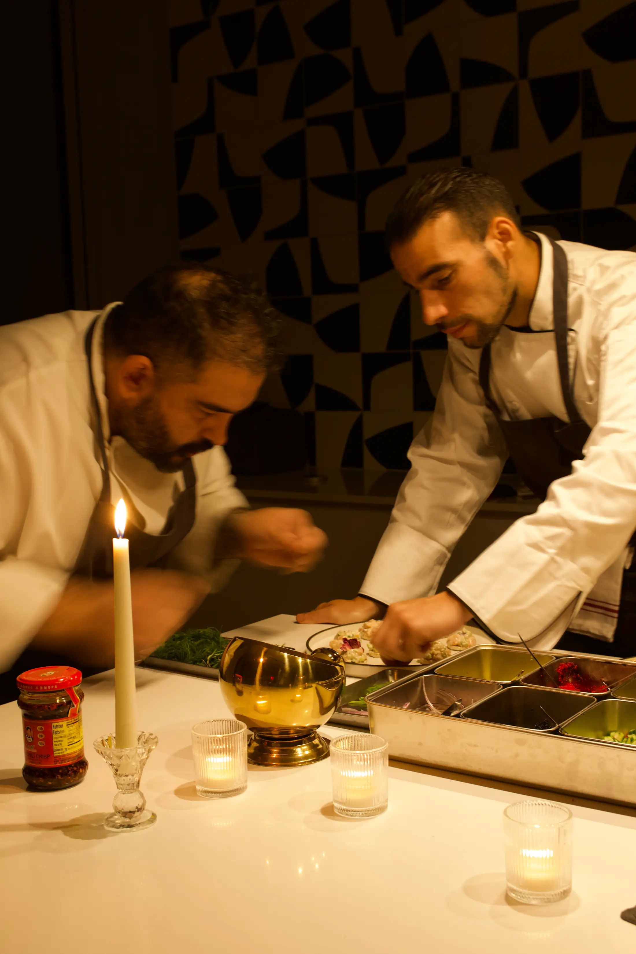 Two chefs in aprons leaning over a counter to finish a plate, candles in front