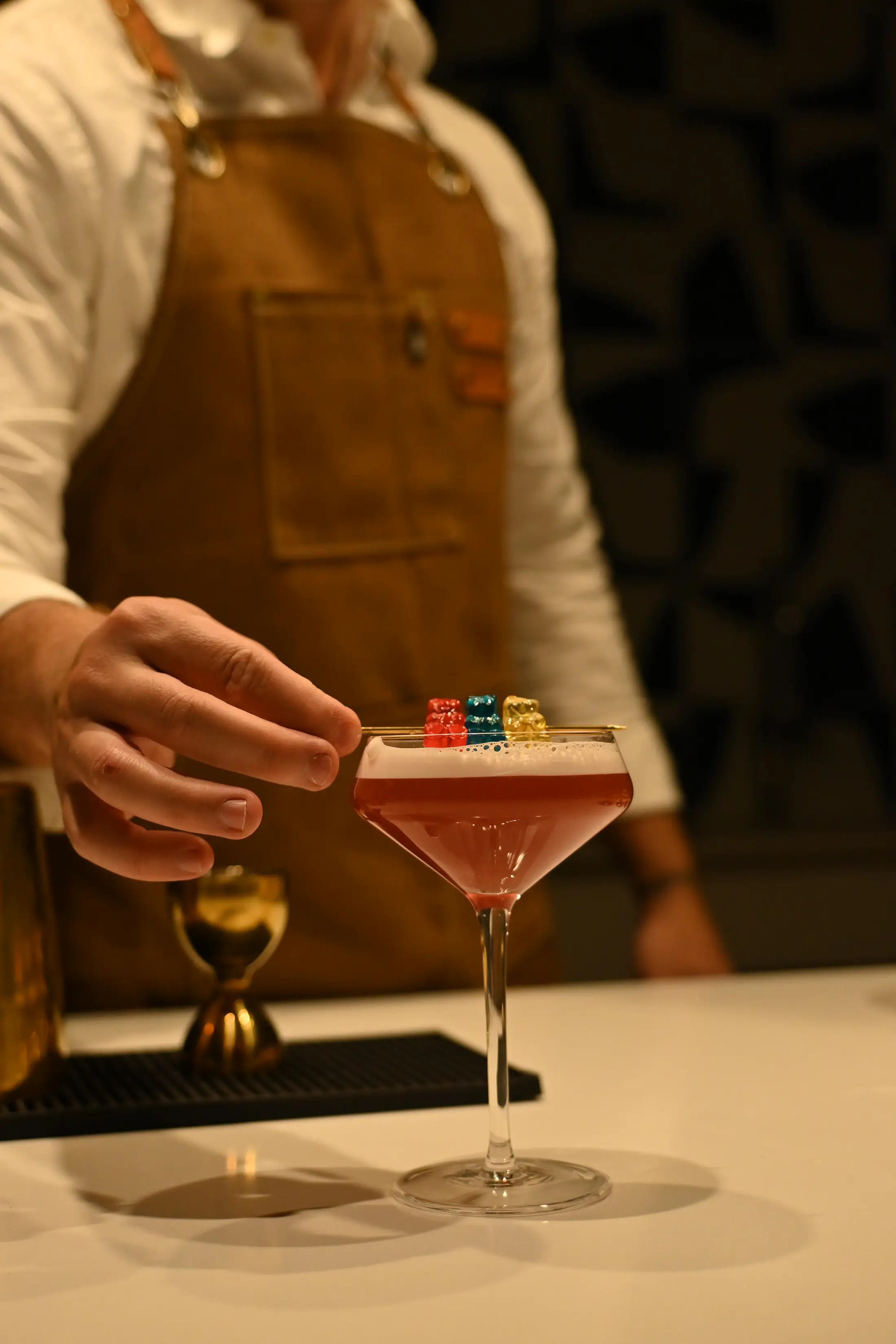 A hand setting a skewer of three gummy bears across a foamed pink cocktail