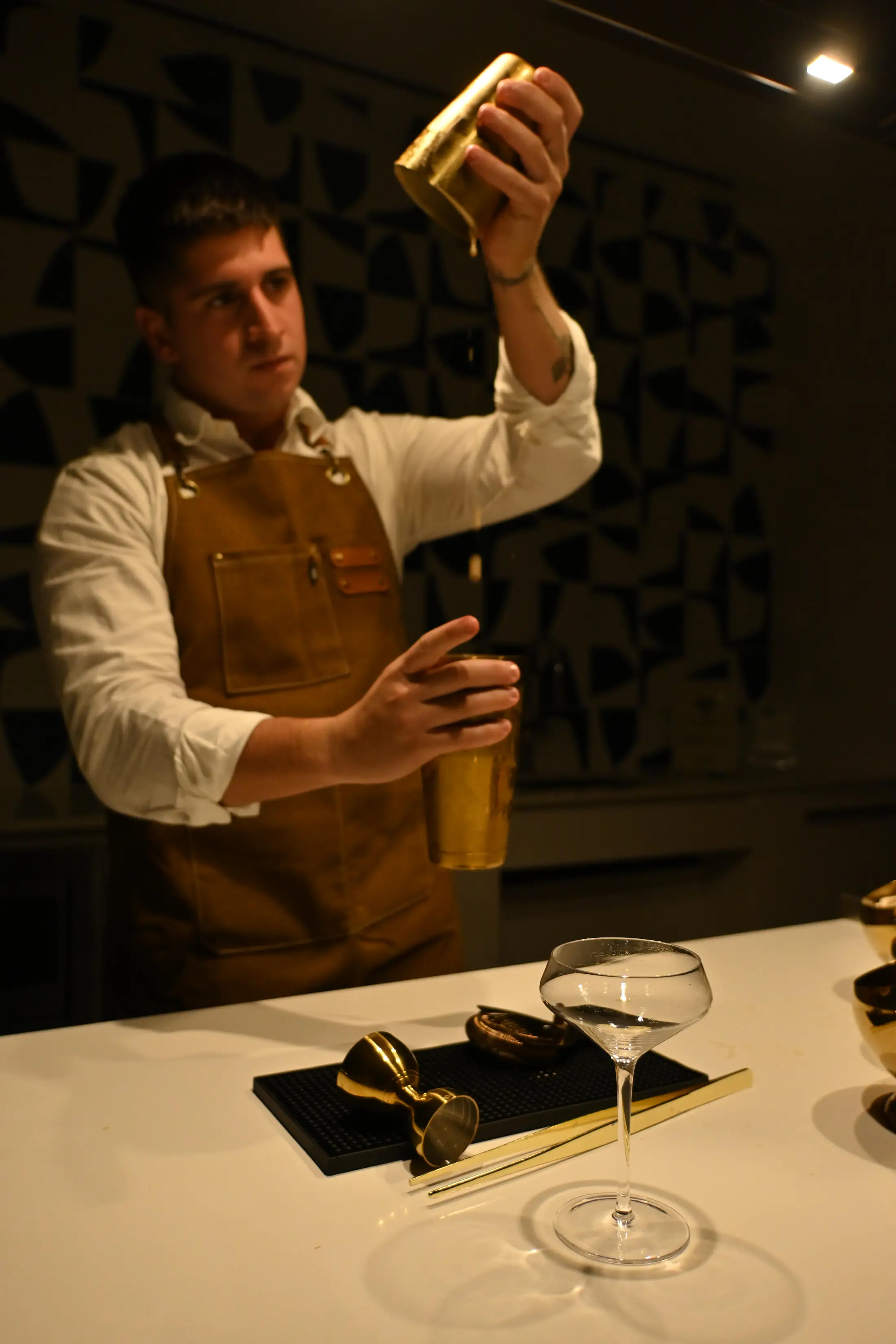A bartender in a leather apron pouring from a gold shaker into a glass