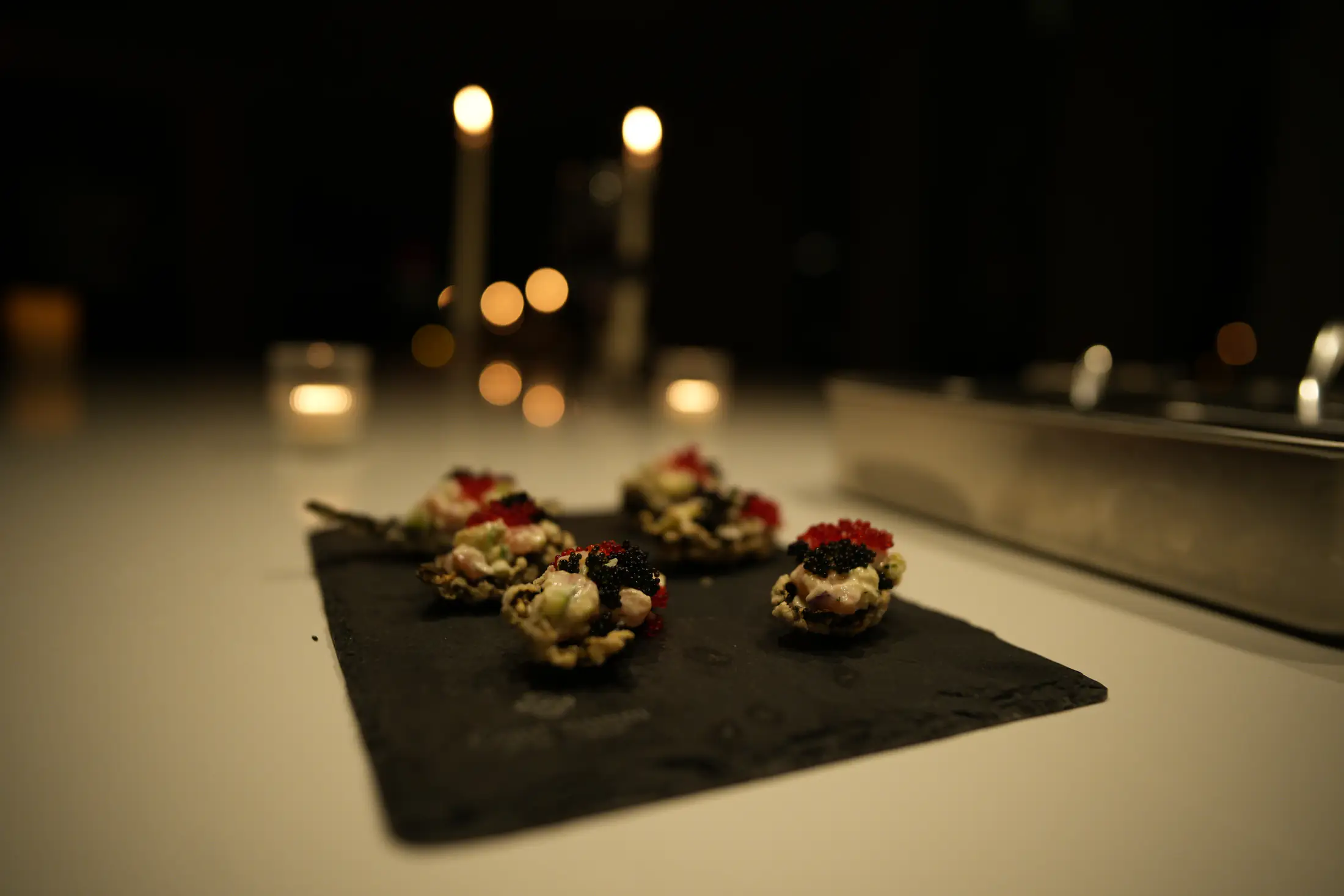 Small bites on a dark slate on the counter, candle flames blurred behind them