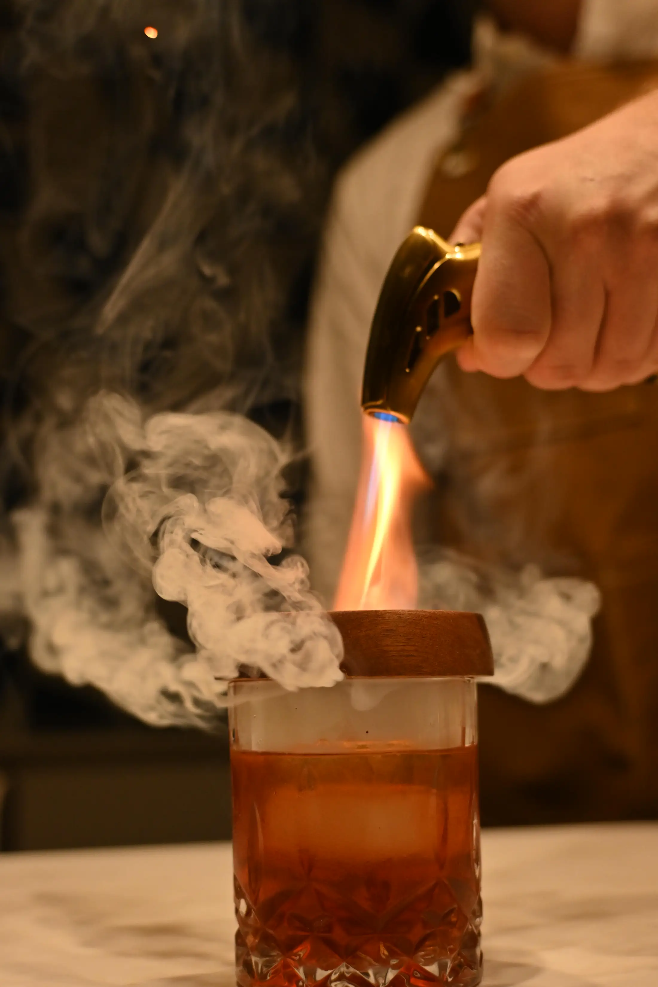 A hand holds a torch to a wooden lid over a smoking amber cocktail