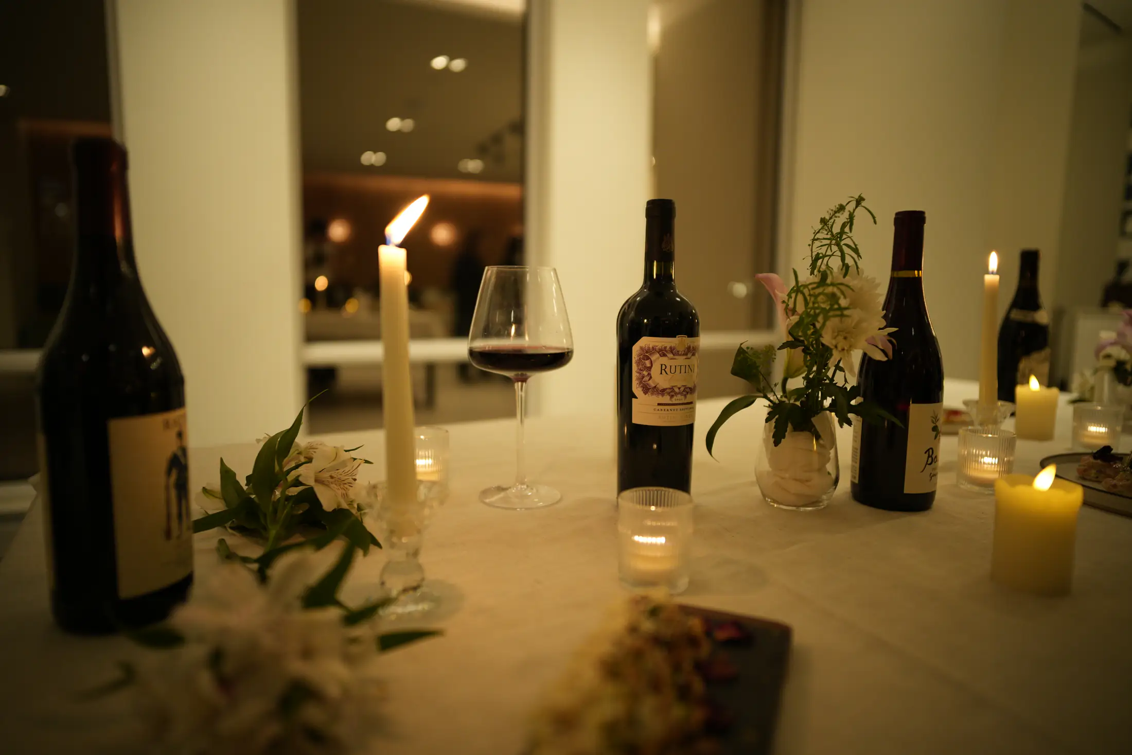 Wine bottles, a poured glass, flowers and lit candles along a white table
